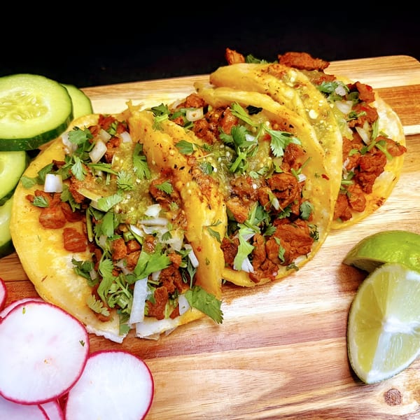 Three Takozz tacos with meat, cilantro, onions, and salsa verde, served with lime, radishes, and cucumbers.