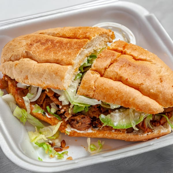 A large Mexican torta sandwich, cut in half, filled with meat, lettuce, and sauce in a takeout container.