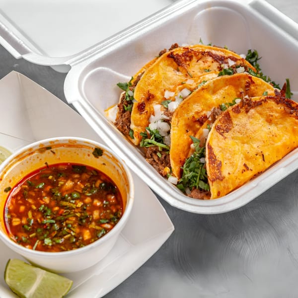 Three birria tacos with cilantro and onions in a takeout container, with a side of consommé and lime.